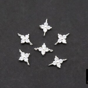 May include: Six silver-coloured, four-petal flower charms with clear, sparkling cubic zirconia stones. Each charm has a small loop for attaching to jewellery. The charms are scattered on a dark grey background.