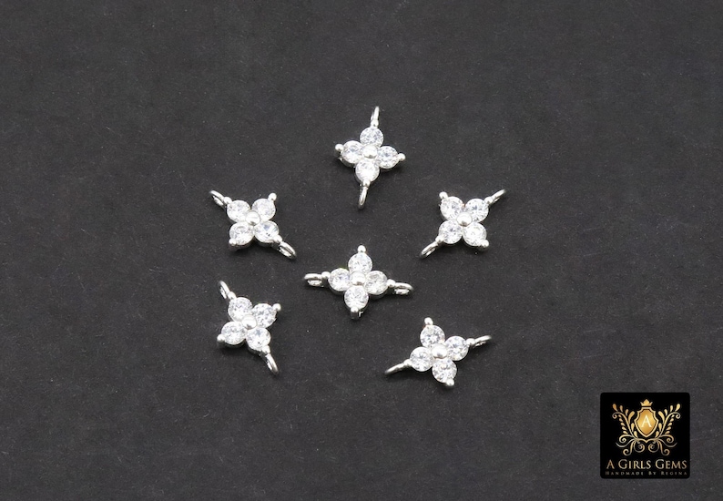 May include: Six silver-coloured, four-petal flower charms with clear, sparkling cubic zirconia stones. Each charm has a small loop for attaching to jewellery. The charms are scattered on a dark grey background.