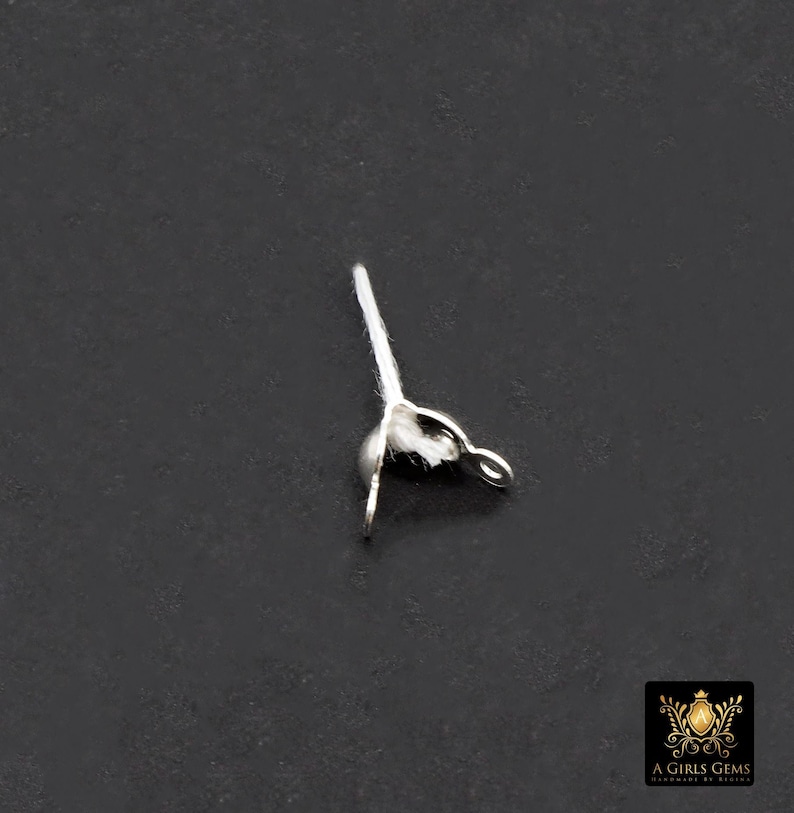 May include: A small, silver-colored jewelry finding with a loop and a small, round bead. A white, fibrous material is attached to the bead. The finding is set against a dark gray background.