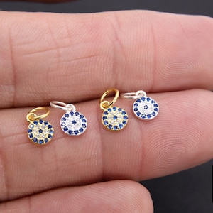 May include: Four silver and gold evil eye charms with blue and white stones. The charms are on a hand.