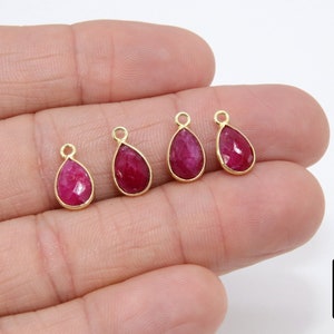 May include: Four gold teardrop-shaped pendants with red gemstone inserts. The pendants are on a hand.