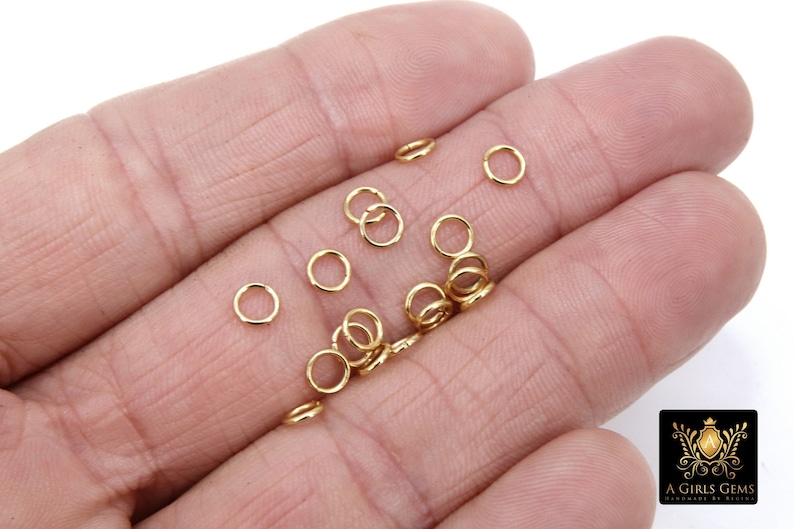 May include: A close-up of a hand holding a group of small, gold jump rings. The rings are about 5mm in diameter and are perfect for jewelry making.