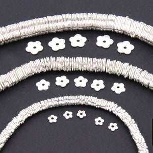 May include: Silver metal beads in various sizes, arranged in a circular pattern. The beads are flat and have a slightly wavy texture. There are also several small silver flower-shaped charms scattered around the beads.