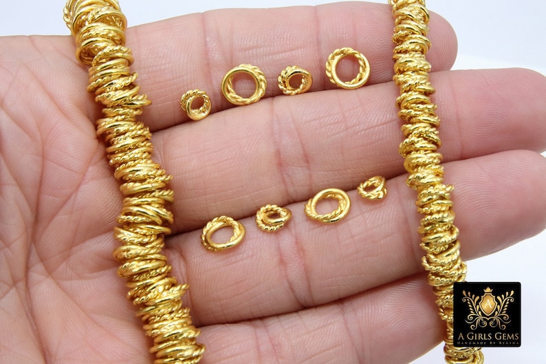 Pu&ograve; includere: Primo piano di una mano che tiene un filo di anelli di metallo dorati. Gli anelli sono piccoli e hanno un design intrecciato. Ci sono anche diversi anelli singoli sparsi sulla mano.