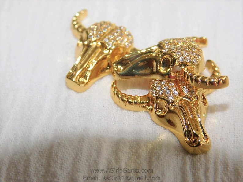 May include: Two gold-tone bull head pendants with clear rhinestones. The pendants are connected by a gold-tone chain.