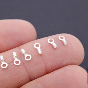 May include: Close-up of six silver crimp beads on a finger. The beads have a small loop on one end and a crimping barrel on the other end.