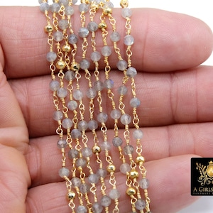 May include: A strand of grey labradorite gemstone beads with gold chain links and gold beads. The strand is held in a hand.