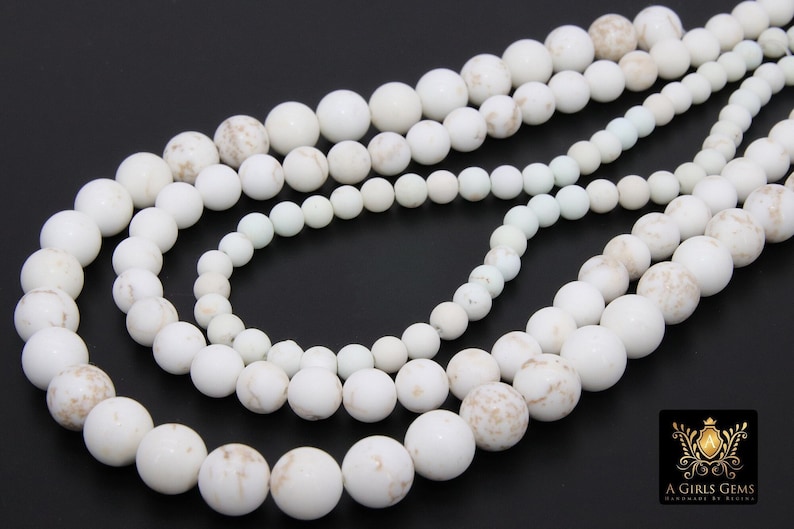 May include: A strand of white howlite beads with brown marbling. The beads are round and smooth, and they are strung on a white cord. The beads are arranged in a circular pattern, and they are all of the same size.