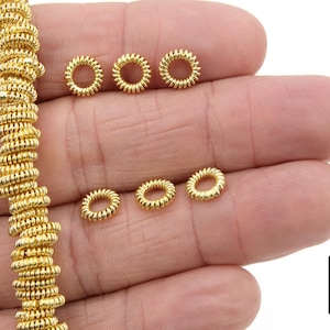 Op de afbeelding: Een close-up van een hand die een streng gouden metalen springringen vasthoudt. De springringen zijn klein en gekruld. Er zijn ook verschillende losse springringen op de hand.