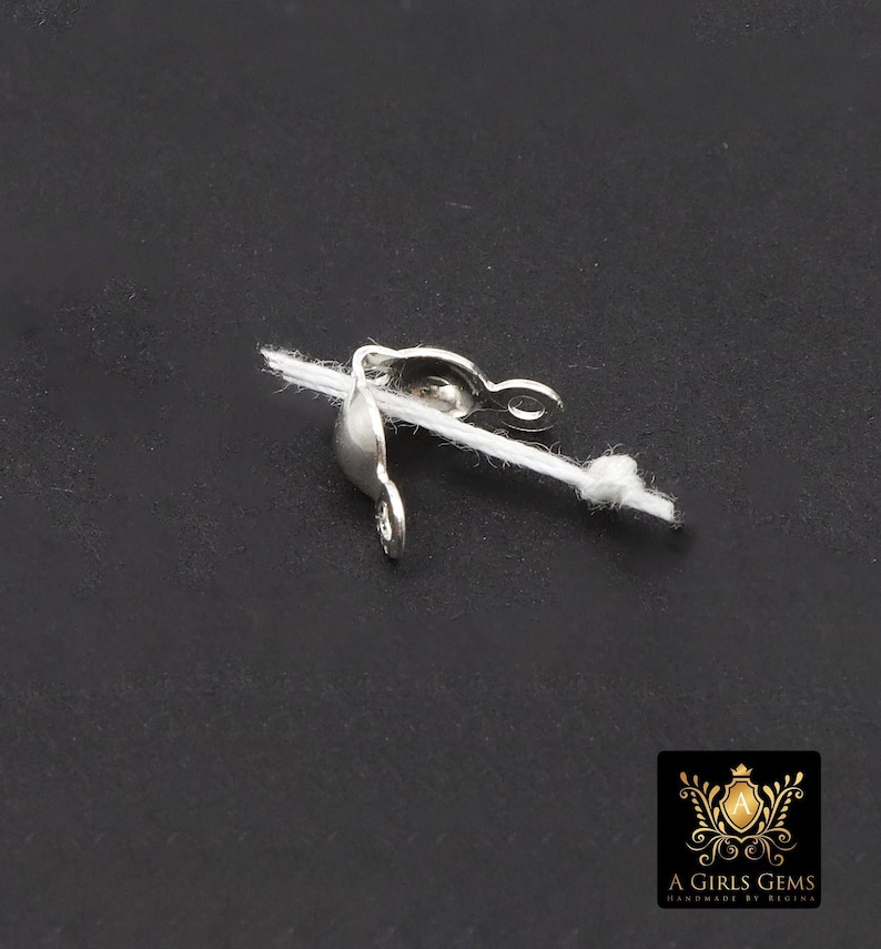 May include: A silver-colored jewelry clasp attached to a white string, set against a dark background. The clasp has a curved design with a loop and a hook. The image also includes a logo in the bottom right corner.
