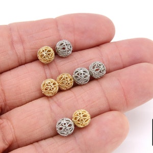 May include: A close-up of six gold and silver filigree beads. The beads are round and have a delicate, intricate design. They are arranged in a hand, with the gold beads on the left and the silver beads on the right.