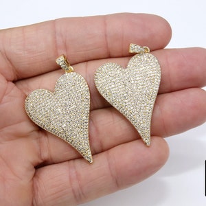 K&ouml;nnte beinhalten: Zwei goldene herzf&ouml;rmige Anh&auml;nger, die mit kleinen, klaren Zirkonia-Steinen besetzt sind. Die Anh&auml;nger befinden sich auf einer Hand, und die Spitzen der Herzen sind spitz.