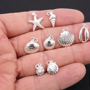 May include: A collection of silver-toned charms featuring seashells and a starfish. The charms are displayed on a hand.