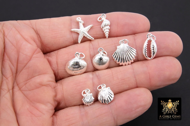 May include: A collection of silver-toned charms featuring seashells and a starfish. The charms are displayed on a hand.