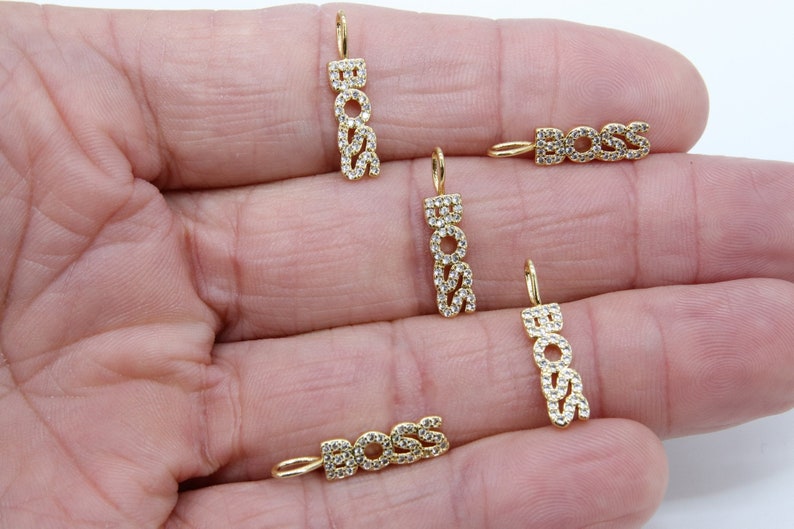 May include: Five gold-tone charms with the word "BOSS" spelled out in clear cubic zirconia stones. The charms are on a hand.