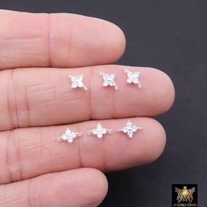May include: Six small, silver-toned flower-shaped earrings with clear, sparkling gemstones are displayed on a hand. The earrings have a four-petal design with a central gemstone. The background is a dark colour.