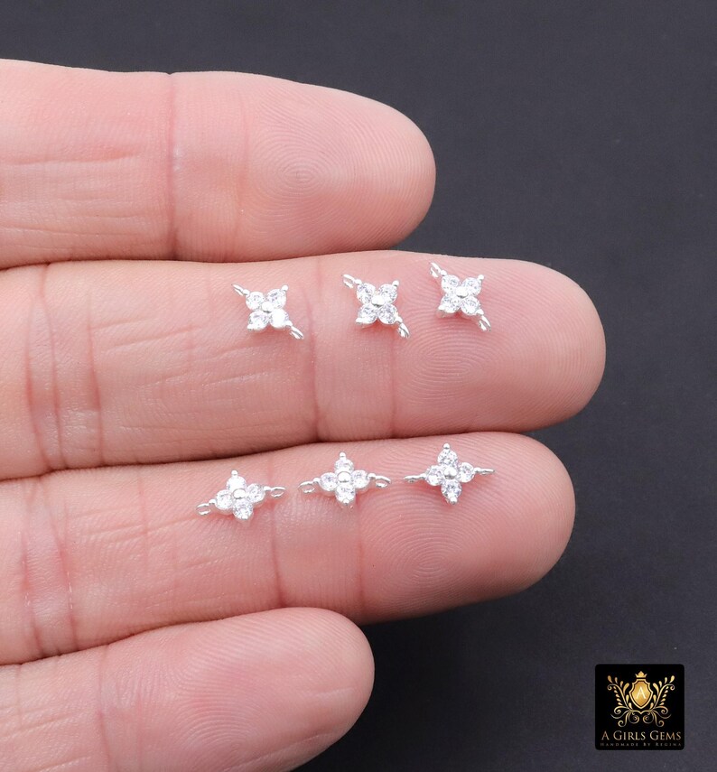 May include: Six small, silver-toned flower-shaped earrings with clear, sparkling gemstones are displayed on a hand. The earrings have a four-petal design with a central gemstone. The background is a dark colour.