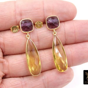 May include: Gold-toned earrings with a square, purple gemstone top and a teardrop-shaped, yellow gemstone pendant. The earrings are held in a hand, showcasing their design and craftsmanship. The earrings are from A Girls Gems.