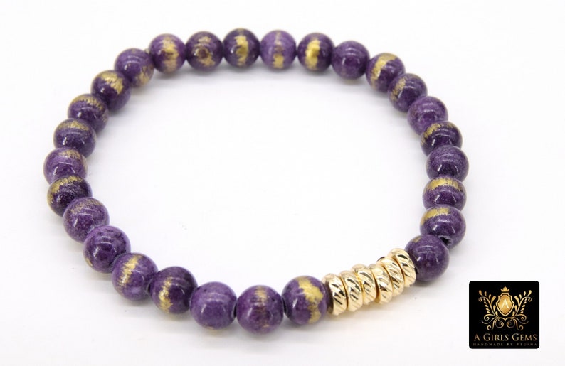 May include: Purple and gold beaded bracelet with a gold accent. The beads are round and have a speckled texture. The bracelet is made by A Girls Gems.