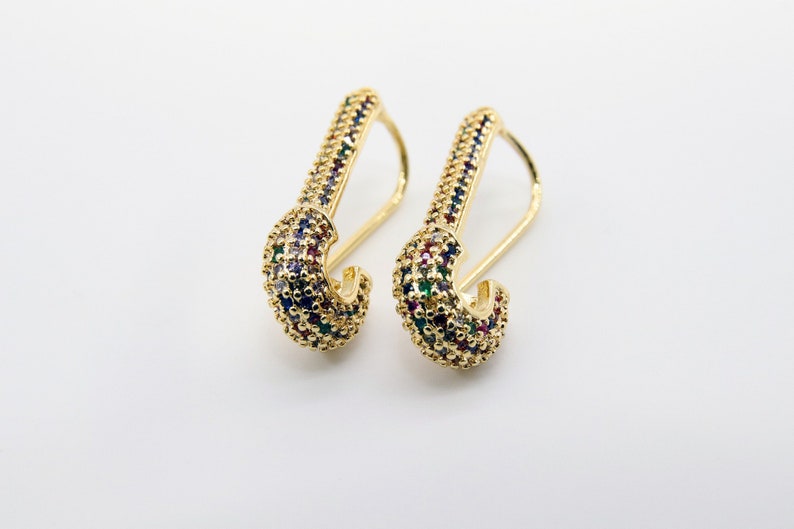 May include: A pair of gold-tone earrings with a safety pin design. The earrings are decorated with colorful cubic zirconia stones.