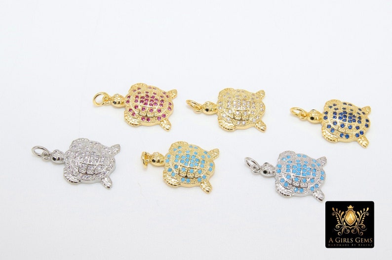 Op de afbeelding: Zes gouden schildpadbedels met verschillende gekleurde kristallen accenten. De bedels zijn bezet met heldere, blauwe, roze en donkerblauwe kristallen.