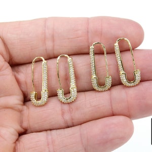 May include: A pair of gold safety pin earrings with clear cubic zirconia stones. The earrings are shown on a hand.