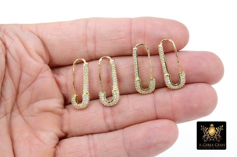 May include: A pair of gold safety pin earrings with clear cubic zirconia stones. The earrings are shown on a hand.