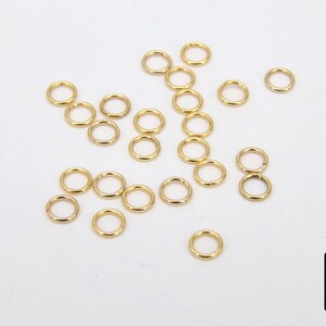 May include: A close-up of a group of 20 gold jump rings. The jump rings are arranged in a random pattern on a white surface.