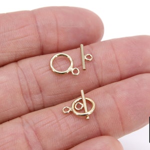 May include: Two gold-toned metal clasps for jewelry making. Each clasp features a round ring and a straight bar with a small loop at one end.