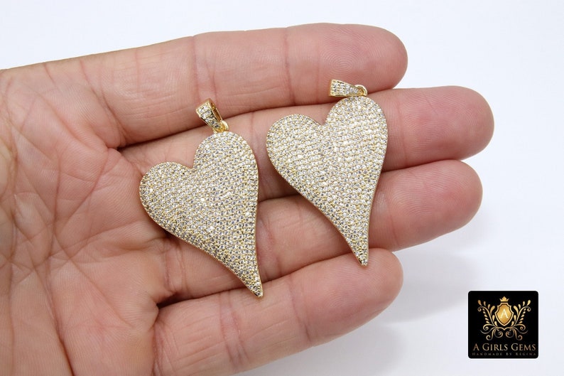 K&ouml;nnte beinhalten: Zwei goldene herzf&ouml;rmige Anh&auml;nger, die mit funkelnden, klaren Steinen besetzt sind. Die Anh&auml;nger haben eine spitze Unterseite und werden in einer Hand gehalten.