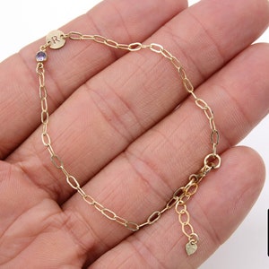May include: A gold chain bracelet with a small purple gemstone and a charm with the letter "R" on it. The bracelet has a heart-shaped charm at the end.