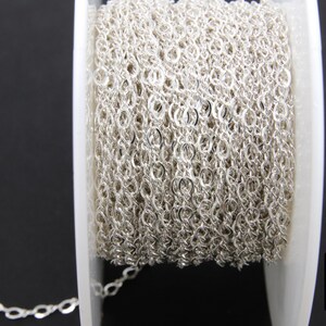 May include: A spool of silver chain, wound tightly, with a small section of the chain extending from the spool. The chain is a delicate, oval-link style.
