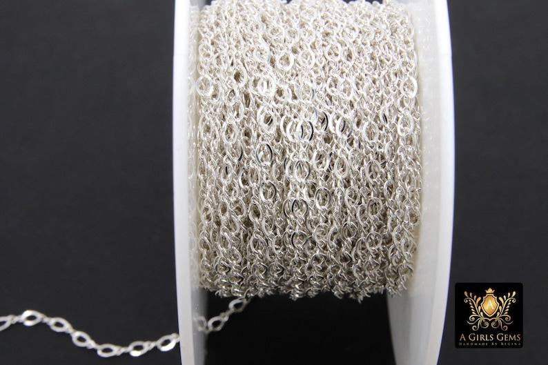 May include: A spool of silver chain, wound tightly, with a small section of the chain extending from the spool. The chain is a delicate, oval-link style.