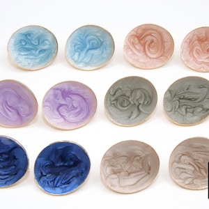 May include: A collection of twelve round, colorful earrings with a swirling, marbled design. The earrings feature shades of blue, pink, purple, gray, and brown, each with a gold-colored edge. The earrings are displayed on a white surface.