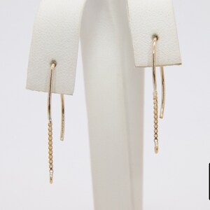 Pu&ograve; includere: Un paio di orecchini pendenti in oro con un delicato design a catena. Gli orecchini sono su un espositore bianco.