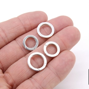 Op de afbeelding: Vier zilveren splitringen, ook wel bekend als jump rings, worden op een hand getoond. De ringen zijn open en klaar om te worden gebruikt voor het bevestigen van bedels, hangers of andere sieradencomponenten.