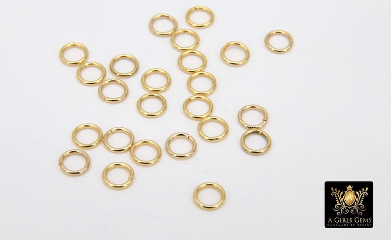 May include: A large quantity of gold jump rings, scattered on a white surface. These rings are commonly used in jewelry making.