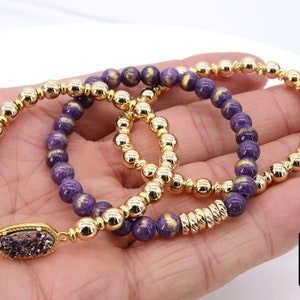 May include: Three gold and purple beaded bracelets with a gold charm featuring a purple and iridescent stone. The bracelets are stacked on a hand.