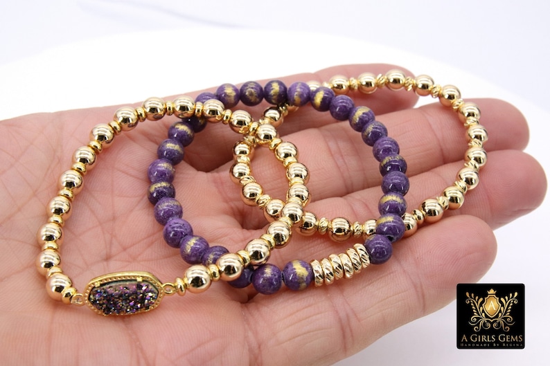May include: Three gold and purple beaded bracelets with a gold charm featuring a purple and iridescent stone. The bracelets are stacked on a hand.