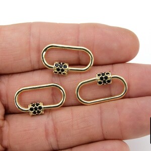 May include: Three gold-colored oval-shaped clasps with black gem accents. The clasps are held in a hand, showcasing their design and detail. The image is well-lit, highlighting the jewelry's features.
