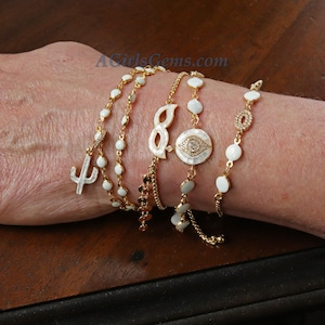 May include: A set of five gold bracelets with white enamel charms. The charms include a cactus, an infinity symbol, an evil eye, and two round beads. The bracelets are adjustable and can be worn together or separately.
