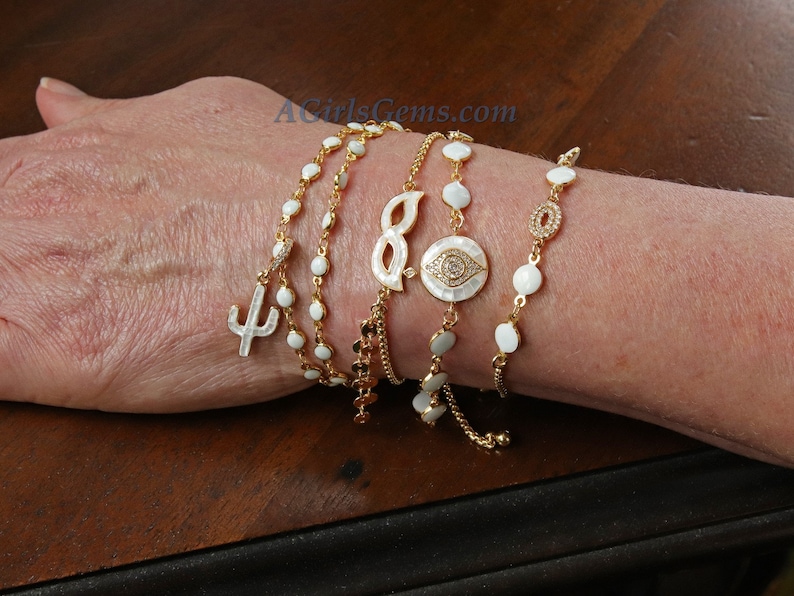 May include: A set of five gold bracelets with white enamel charms. The charms include a cactus, an infinity symbol, an evil eye, and two round beads. The bracelets are adjustable and can be worn together or separately.