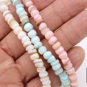 May include: Strands of colorful gemstone beads in pastel shades of white, blue, and pink. The beads are small and have a rounded shape. The image is of a hand holding the strands of beads. The logo "A Girls Gems" is in the bottom right corner.