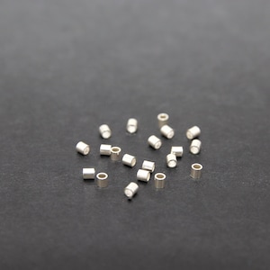 Pode incluir: Uma coleção de pequenas contas de crimpagem de metal prateado espalhadas sobre uma superfície escura. Estas contas cilíndricas têm um orifício que atravessa o centro e são usadas na fabricação de joias para prender fechos e outros componentes.