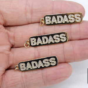 May include: Three gold-tone pendants with the word "BADASS" spelled out in white rhinestones. The pendants are on a black background.