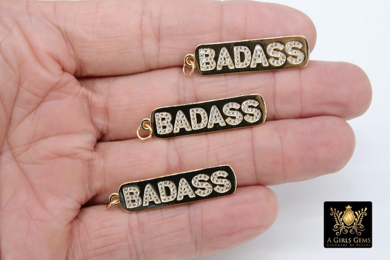 May include: Three gold-tone pendants with the word "BADASS" spelled out in white rhinestones. The pendants are on a black background.
