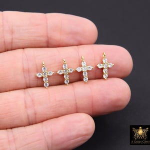 May include: Four small gold-colored cross pendants, each adorned with clear, sparkling gemstones. The crosses are displayed against a neutral background, showcasing their intricate design and craftsmanship. The pendants are delicate and suitable for jewelry making.