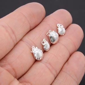 May include: Four silver-toned seashell charms with a loop for attaching to jewelry. The charms are arranged in a row on a hand.