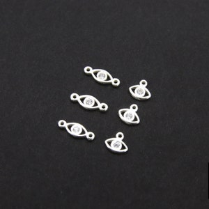 May include: Six silver eye charm pendants with clear rhinestone accents. The charms are oval shaped with a small loop at the top for attaching to jewelry.