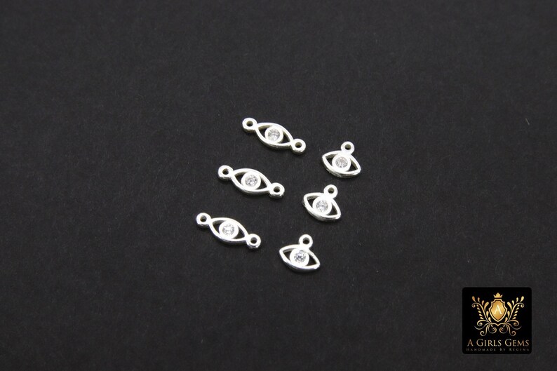 May include: Six silver eye charm pendants with clear rhinestone accents. The charms are oval shaped with a small loop at the top for attaching to jewelry.